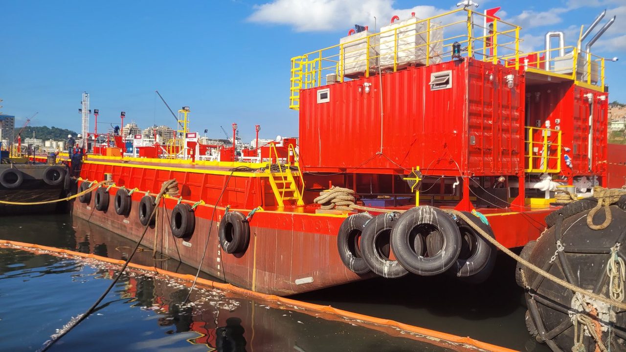 Balsa de apoio atracada em estaleiro, com módulos containerizados, guarda-corpos e defensas, durante inspeção naval.