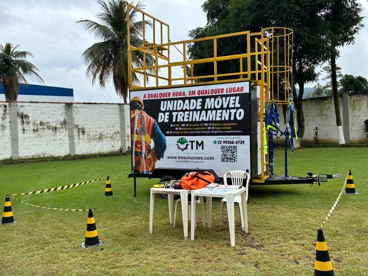 Unidade móvel de treinamento da TM montada em área externa, com guarda-corpo superior para prática de trabalho em altura, cintos paraquedistas e área isolada por cones e fita zebrada.
